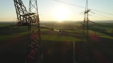 Gün batımında boş yeşil ve sarı alanlar arasında gökyüzüne karşı elektrik desteklerinin videosunu kapat. Medeniyet arka planda görülebilir. Güneşli gün, yaz, kırsal bölge, yüksek voltaj hattı