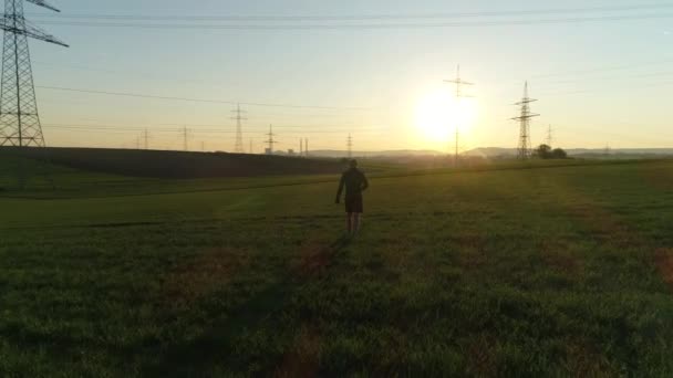 Kale man in het zwart loopt op het veld bij zonsondergang. Elektrische steunen tegen de lucht tussen lege groene en gele velden. Zonnige dag, zomer, platteland, hoogspanningslijn. 4K-video. — Stockvideo