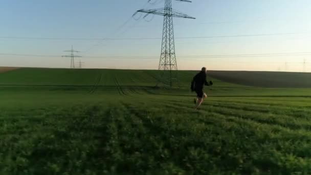 Un hombre calvo de negro corre por el campo al atardecer. Soportes eléctricos contra el cielo entre campos verdes y amarillos vacíos. Día soleado, verano, campo, línea de alto voltaje. Vídeo 4K. — Vídeos de Stock