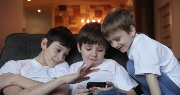Chicos sentados en el sofá y jugar juegos en línea. Los jóvenes usan sus teléfonos. tres niños en camisetas blancas. — Vídeo de stock
