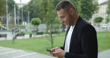 Caddenin yanında yürüyen genç işadamının yakınında. Akıllı Telefonun Ekranı 'na bakıyorum. Klasik giyiniklik. Arka plandaki Büyük Cam Bina. Başarılı ve kendinden emin görünüyor..