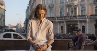 Çekici bir adam dışarıda oturuyor ve akıllı telefon kullanıyor. Genç adam akıllı telefona bakarken gülüyor. Mutluluk duygusu