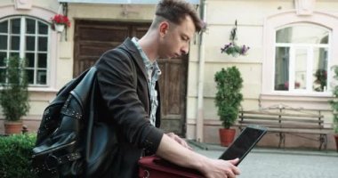 Ceketli, siyah sırt çantalı, şık saç kesimi olan çekici bir adam dışarıda dizüstü bilgisayarla çalışıyor. Öğrenci.