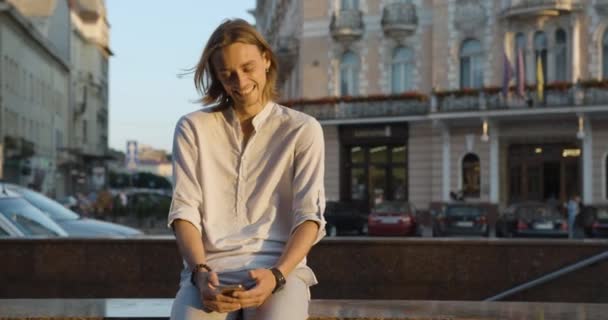 Un chico atractivo se sienta fuera y utiliza un teléfono inteligente. joven riendo mientras mira el teléfono inteligente. emoción de felicidad — Vídeos de Stock