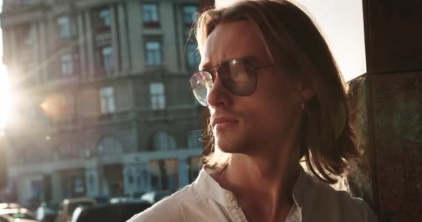 Retrato de primer plano de un joven atractivo en gafas de pie en la calle contra el fondo del atardecer y el edificio de gran altura. — Vídeos de Stock