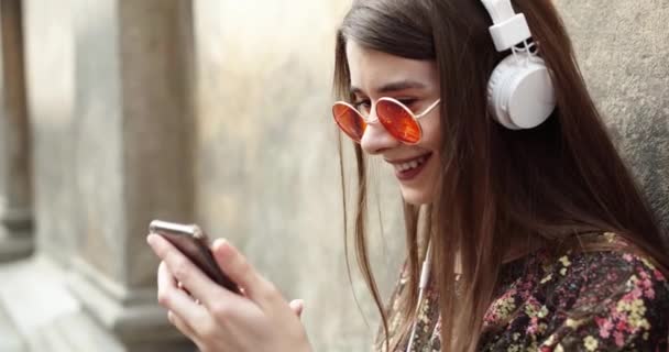 Ritratto di una ragazza felice che ascolta musica online con cuffie wireless da smartphone in strada in una giornata di sole estivo. ragazza con le cuffie bianche. ragazza in bicchieri rotondi rossi — Video Stock