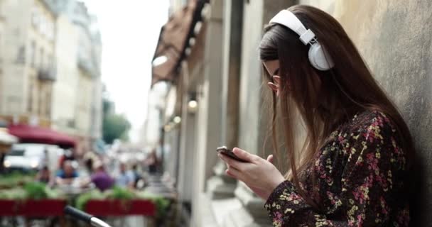 Menina atraente em fones de ouvido com smartphone ouvindo música na rua. menina bonito com fones de ouvido brancos e óculos redondos vermelhos — Vídeo de Stock