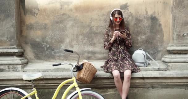 Fiatal kaukázusi barna nő fejhallgatóval a szabadban, napsütéses nyári napon. Millenniumi tinédzser lány zenét hallgat. Négyzet alakú, retusált, szűrő. Sárga kerékpár. — Stock videók
