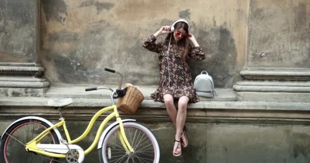 Piękna dziewczyna z długimi włosami siedząca na ulicy i słuchająca muzyki w białych gorących telefonach czerwone okulary przeciwsłoneczne i uśmiechnięta. Nastrój muzyczny. Szczęśliwych emocji. Dziewczyna z żółtym rowerem. — Wideo stockowe