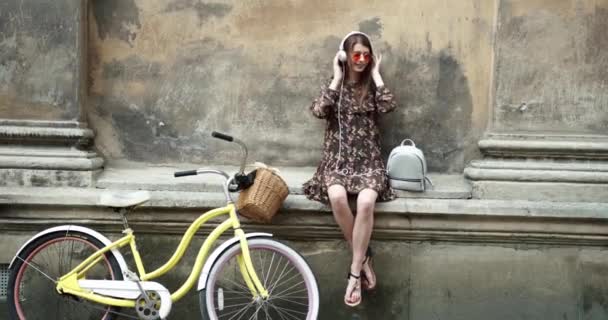 Hermosa chica con el pelo largo sentado en la calle y escuchando música en calentadores blancos gafas de sol rojas y sonrientes. Humor musical. Feliz emoción. Chica con bicicleta amarilla — Vídeo de stock