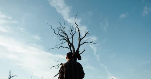 Bakifrån av senior skogshuggare i naturen håller en yxa på axeln och närmar sig ett stort torrt träd. — Stockvideo