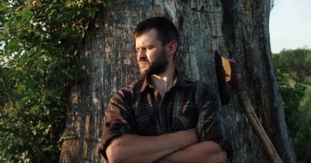Atrakcyjny brodaty mężczyzna w koszuli złożył ręce przed sobą i stoi opierając się o drzewo. siekiera na drzewie — Wideo stockowe
