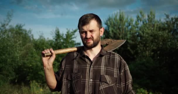 Bonito morena homem em uma camisa fica no meio de uma floresta com um machado em suas mãos. — Vídeo de Stock