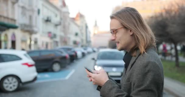 Homem atraente em roupas elegantes passear pela cidade usa um smartphone. o homem parece alegre — Vídeo de Stock