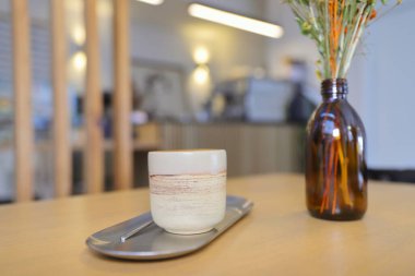 Bir kafe içinde sıcak ve davetkar bir sahne, seramik bir fincanda kremalı latte ve yanında rahatlama ve tefekkür için mükemmel kurutulmuş botanik vazosu.