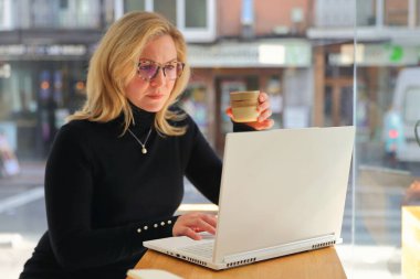 Şık gözlüklü ve siyah balıkçı yaka yakalı bir kadın akıllı telefonuna dalmış, önünde dizüstü bilgisayar açık bir şekilde duruyor, bir çalışma ya da çalışma oturumu öneriyor.