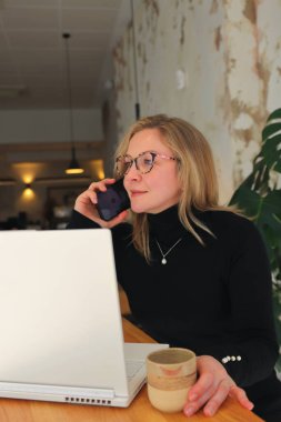 Şık gözlüklü ve siyah balıkçı yaka yakalı bir kadın akıllı telefonuna dalmış, önünde dizüstü bilgisayar açık bir şekilde duruyor, bir çalışma ya da çalışma oturumu öneriyor.