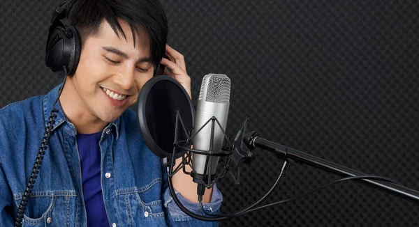 Musician producing music in professional recording studio. Young asian man with headphone singing in front of black soundproof wall.