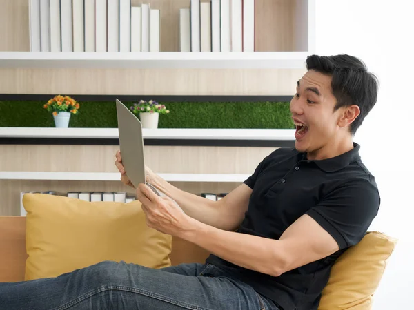 The young man wore comfortable clothes makes a happy expression while reading an e-mail from laptop computer. Asian man spend holidays in the living room.