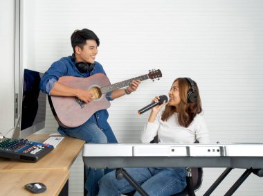 Genç Asyalı kadın beyaz duvarların önünde elektrikli klavye çalarken şarkı söylüyor. Erkek arkadaşı birlikte gitar çalıyordu. Müzisyenler profesyonel kayıt stüdyosunda müzik yapıyor..