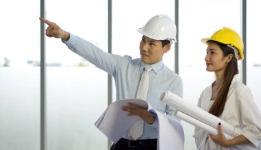 Proje sahibi ve yardımcısı inşaat halindeki projeleri kontrol etmek için koruyucu şapkalar takıyorlar. İnşaat çizimleri kadının elinde..
