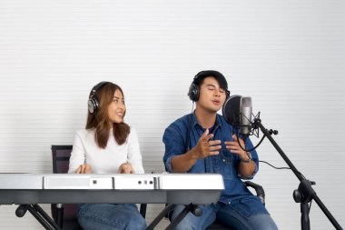 Kulaklıklı genç Asyalı adam beyaz duvarların önünde mikrofonla şarkı söylüyor. Kız arkadaşı birlikte elektrikli klavye çaldı. Müzisyenler profesyonel kayıt stüdyosunda müzik yapıyor..