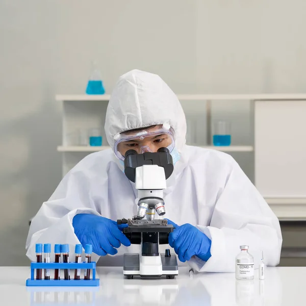 Virüse karşı koruyucu giysi araştırmacısı mikrop kapmış insanların kan örneklerine mikroskop kullanarak bakıyor. Coronavirus hastalığı 2019 (COVID-19) laboratuvarda test süreci.