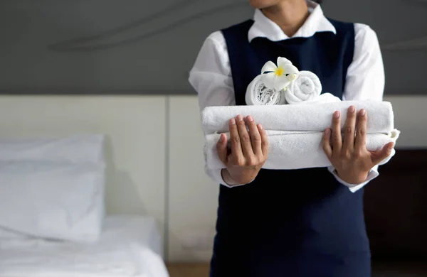 Genç otel hizmetçisinin elinde beyaz çiçekli yakın plan beyaz havlu. Mavi üniformalı personel otel misafirleri için oda hazırlıyor..