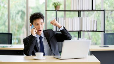 Genç adam dizüstü bilgisayarda e-posta okurken telefonla konuşuyor. Takım elbiseli Asyalı işadamı iyi haberlerden heyecanlanmış, ofiste dizüstü bilgisayarla çalışıyor. (evet jest)