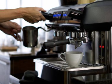 Kahve makinesine beyaz bir seramik bardak yerleştirildi. Espresso kahvesi yapmaya hazırlan. Barista arka planda süt köpürten sürahi tutuyor..