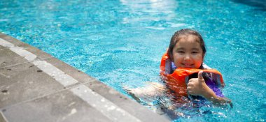 Asyalı çocuk havuzda oynuyor. Turuncu can yeleği giyiyor, başparmağıyla gülümsüyor..