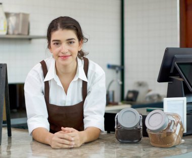 Kahverengi önlük giymiş Kafkasyalı genç kafe çalışanları müşterilerin dükkana girmesini beklerken tezgaha yaslanıyor. Bir kafede sabah atmosferi.