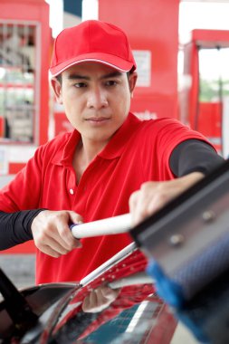 Kırmızı üniformalı Asyalı bir benzin istasyonu çalışanı cam sileceğiyle arabanın ön camını temizliyor..