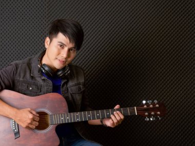 Siyah ses geçirmez duvarların önünde akustik gitar çalan kulaklıklı genç Asyalı bir adam. Müzisyenler profesyonel kayıt stüdyosunda müzik yapıyor..