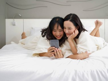 Bornozlu iki Asyalı kadın yatakta yatarken cep telefonuyla selfie çekiyorlar..