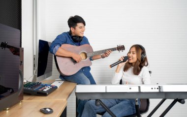 Genç Asyalı kadın beyaz duvarların önünde elektrikli klavye çalarken şarkı söylüyor. Erkek arkadaşı birlikte gitar çalıyordu. Müzisyenler profesyonel kayıt stüdyosunda müzik yapıyor..