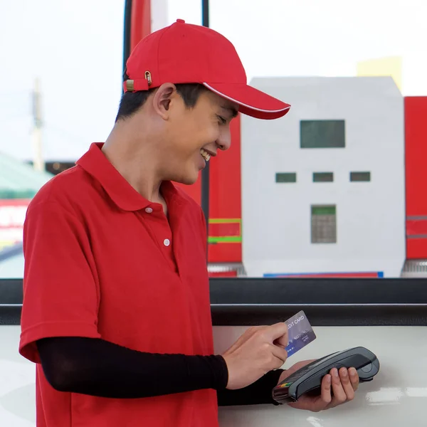 Gas station worker in red uniform stand smiling, swipe mockup credit card via payment terminal. Cash, price and volume readout display on petrol pump display screen.