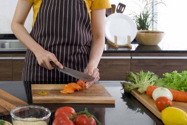 Genç ev hanımı, tahta kesme tahtasıyla kırmızı domatesi parçalara ayırdı. Mutfak tezgahı çeşitli sebzelerle dolu. Modern bir mutfakta sabah atmosferi.