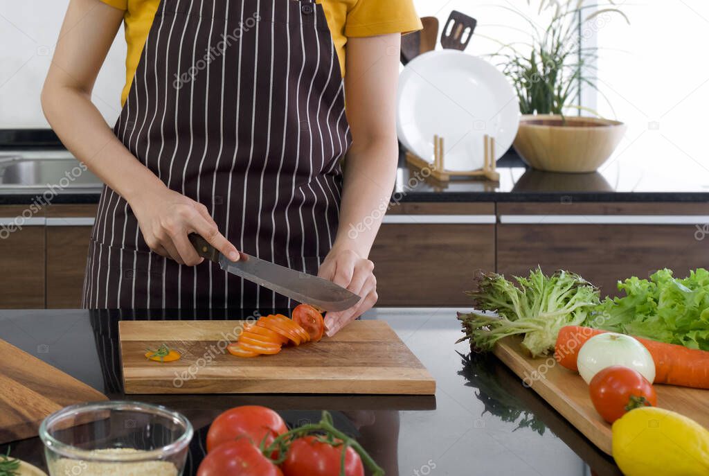 Young housewife slice red tomato into pieces on a wooden chopping board. The kitchen counter full of various kinds of vegetables. Morning atmosphere in a modern kitchen.