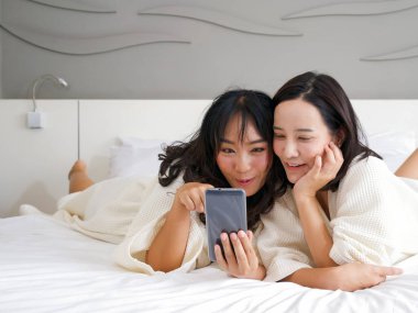 Bornozlu iki Asyalı kadın yatakta yatarken cep telefonuyla selfie çekiyor..