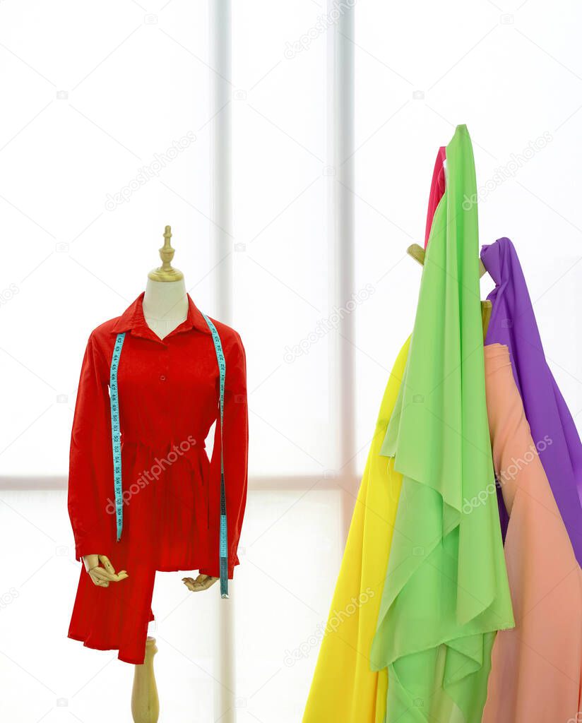 The red shirt and tape measure are on the mannequin. A clothes rack with colorful fabrics in front of the large window. Morning atmosphere in the tailor's shop.