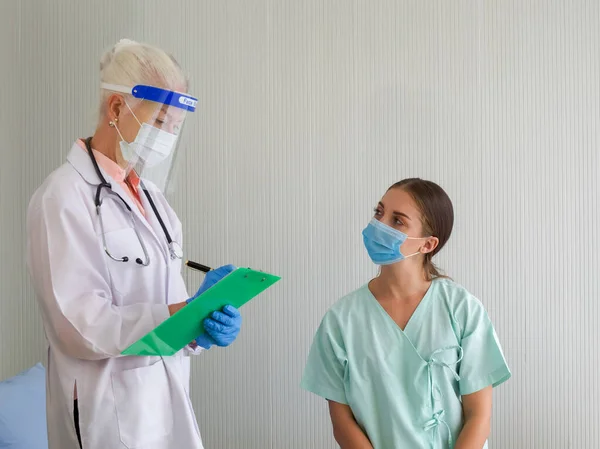 顔シールドと保護マスクを持つ老婦人医師は ベッドに座っている患者の症状について尋ねました 病院でのCovid 19試験 — ストック写真