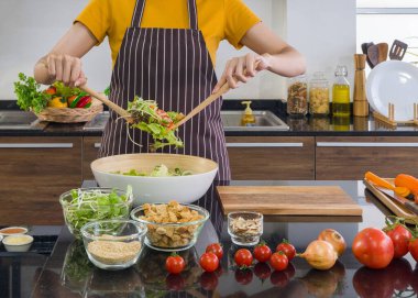 Ev hanımı önlük giymiş, sebzeleri ahşap bir kepçeyle salata kasesine karıştırmış. Modern bir mutfakta sabah atmosferi.