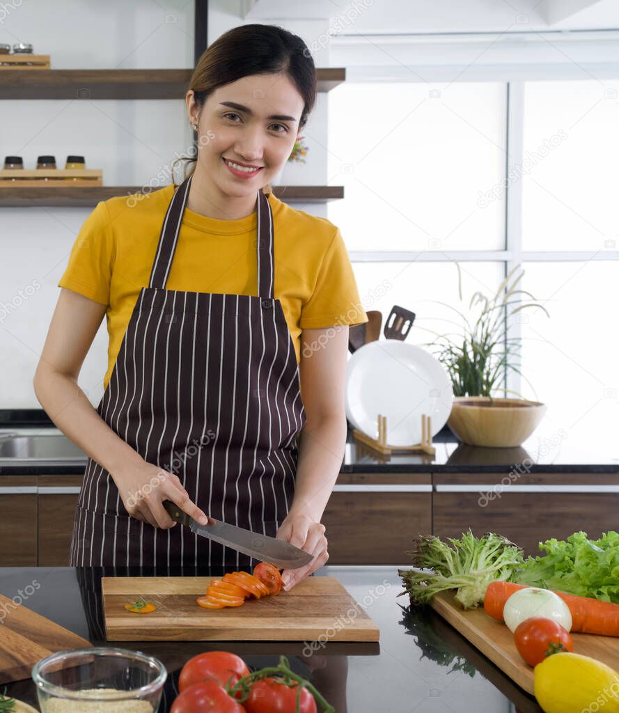 Young housewife slice red tomato into pieces on a wooden chopping board. The kitchen counter full of various kinds of vegetables. Morning atmosphere in a modern kitchen.