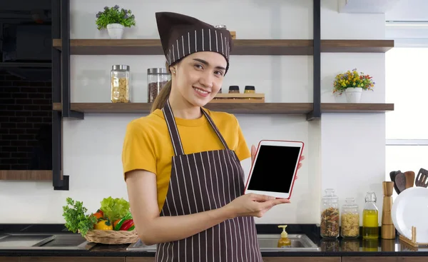 The housewife dressed in an apron and a hair cap, holding blank screen tablet computer. Morning atmosphere in a modern kitchen.