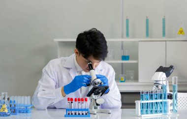 Genç Asyalı bilim adamı hastanın kanını görmek için mikroskop kullanıyor. Coronavirus hastalığı 2019 (COVID-19) viral araştırmaların yayılmasını önleyen bir laboratuvarda test süreci.