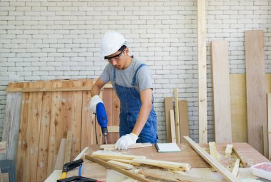 Asyalı marangozlar ahşap fabrikasında hazırlandıkları gibi kalasları delmek için elektrikli matkap kullanıyorlar. Atölyede sabah çalışma ortamı. El aletleri ve odun yığınlarıyla dolu bir masa..