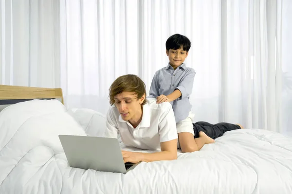 Father work at home with son. The boy ride on his father\'s back while the father using laptop computer to work in the bedroom. Work life balance.