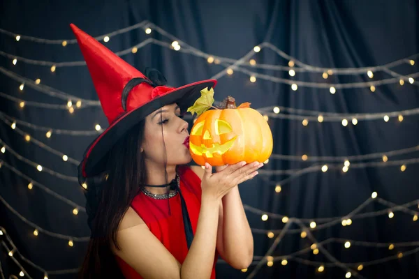 Caucasian woman in red dress  kissing pumpkin carved into a face stand in front of black curtain decoration with lighting at halloween party.