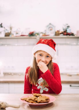 Kırmızı kazak ve Noel Baba şapkasıyla mutfakta zencefilli kurabiye yiyen bir kızın dikey portresi. Metin için yer.